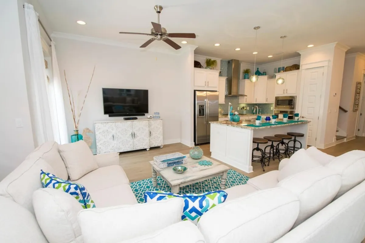 This image showcases a spacious, open-concept living area that flows seamlessly into the kitchen and dining space. The scene features a plush sectional sofa facing a large television, a kitchen island with bar seating, and a dining area visible in the background. The room is filled with natural light and decorated in a welcoming, modern style.