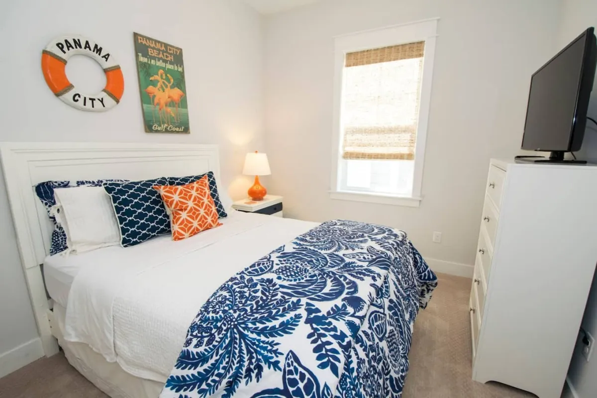 A bright bedroom featuring a bed with blue and white bedding, an orange accent pillow, a nightstand with a lamp, framed wall art, and a mounted flat-screen television. The space feels welcoming and cheerful, ideal for rest and relaxation.