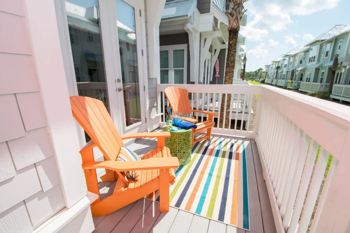 The image showcases a cheerful outdoor seating area at the front of the beach house, featuring two bright orange Adirondack chairs atop a striped, colorful outdoor rug. A small green side table holds drinks and accessories, inviting guests to relax and soak up the Florida sunshine. The setup is perfect for unwinding with a cold beverage, reading, or enjoying lively conversation, all set against the relaxed coastal backdrop of 30A.