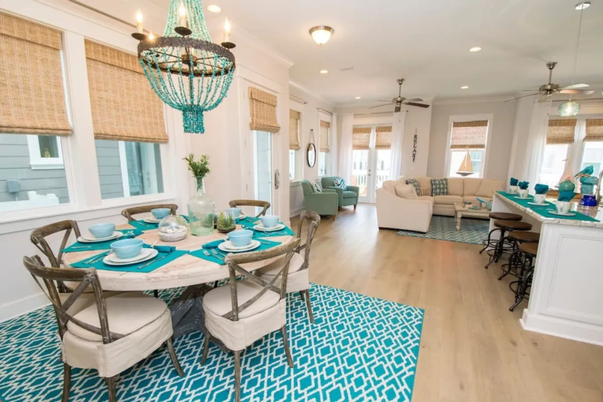 A bright, open-plan kitchen and dining area featuring a turquoise beaded chandelier, a dining table set with turquoise dinnerware, a comfortable sectional sofa, and a kitchen island with bar stools. The space is welcoming and designed for both cooking and socializing.