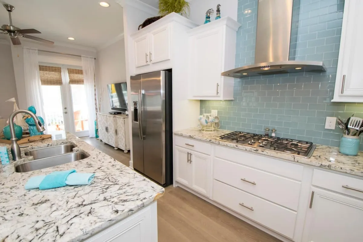 A modern kitchen scene with marble countertops, a stainless steel refrigerator, a blue subway tile backsplash, and a sleek gas cooktop and hood. The kitchen radiates freshness and sophistication, with custom finishes and high-end appliances.