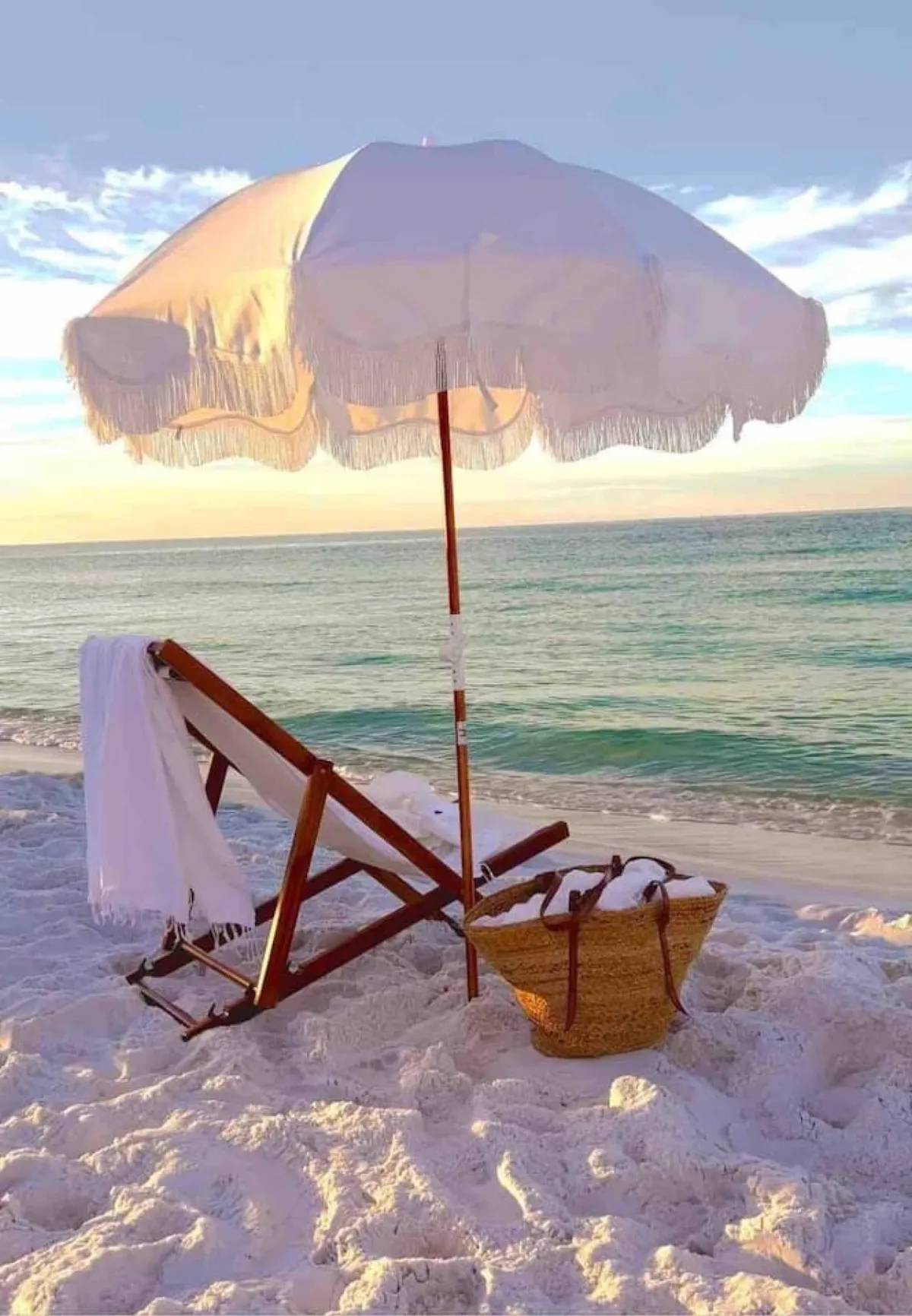 A classic wooden beach chair with white fabric sits beneath a large, fringed white umbrella on the sandy beach. A woven basket rests nearby, and gentle ocean waves form a tranquil backdrop, suggesting a peaceful, well-equipped setting for beach relaxation.