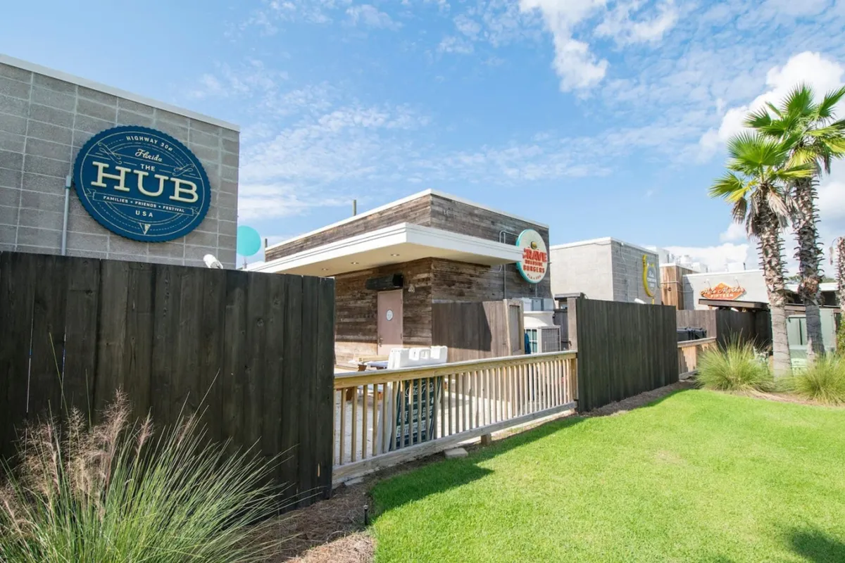 The Hub sign marks a rustic wood-paneled building with a lively outdoor seating area, palm trees, and a green lawn, suggesting a vibrant communal hub for dining, concerts, or movie screenings. The setting highlights outdoor leisure and community engagement.