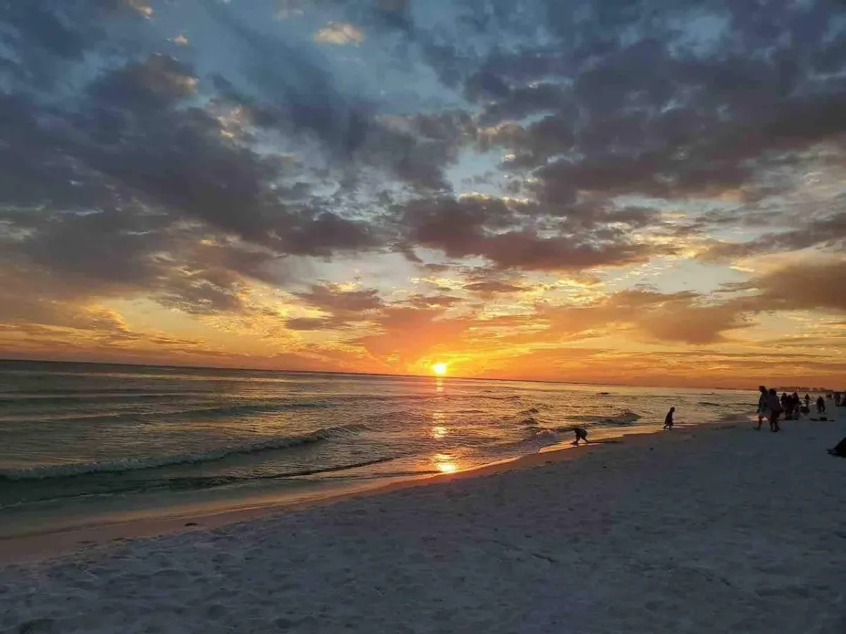 A serene sunset over the beach, with people—likely families—walking, children playing in the sand and water, and gentle waves washing ashore. The soft, reflective light captures both relaxation and lively family enjoyment on the sand.