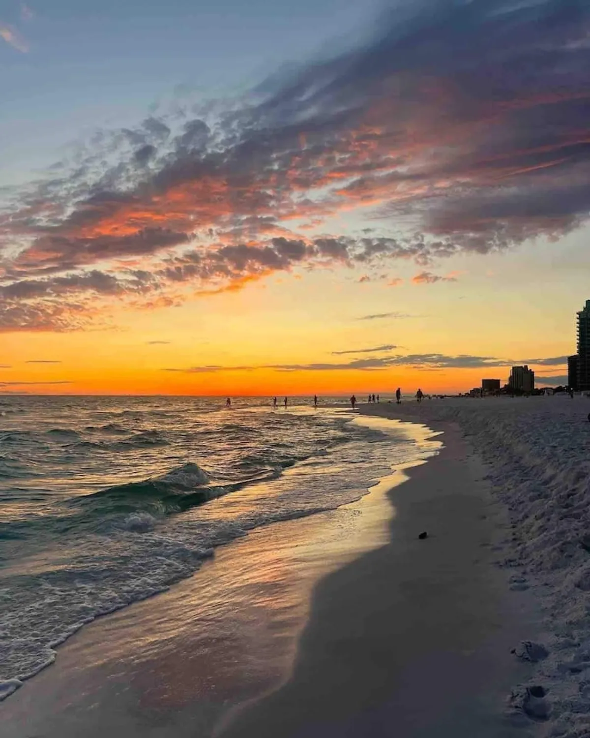 A vivid sunset washes the sky in orange and purple hues as gentle waves lap the sandy shore. Silhouetted people stroll along the waterline, with beachfront buildings faintly visible on the horizon, evoking a sense of local community and relaxation.
