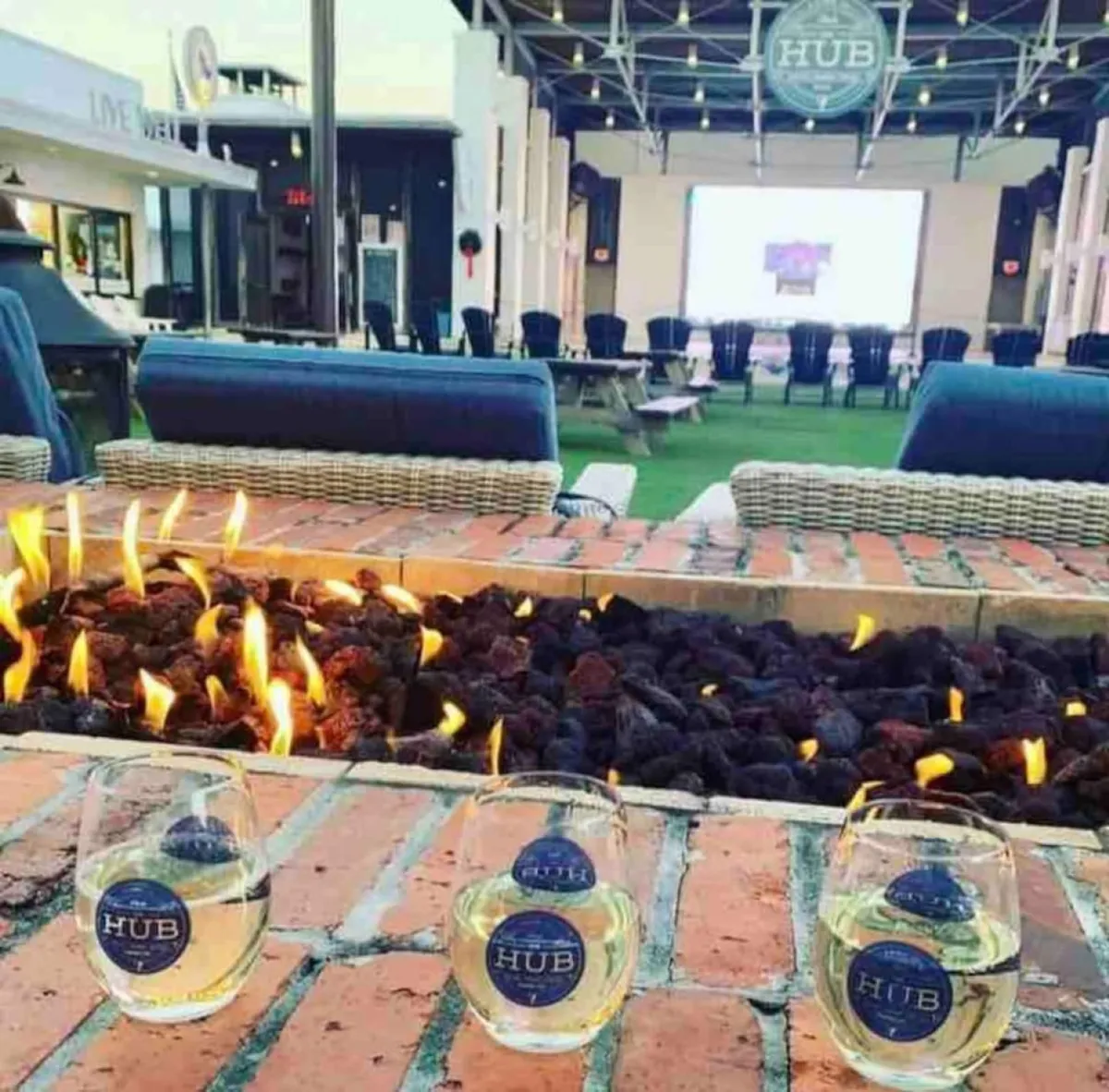 An inviting brick fire pit with glowing flames is surrounded by comfortable outdoor seating with blue cushions and a backdrop of an amphitheater screen, where guests enjoy drinks and socialize. The lively scene reflects both communal entertainment and upscale amenities.