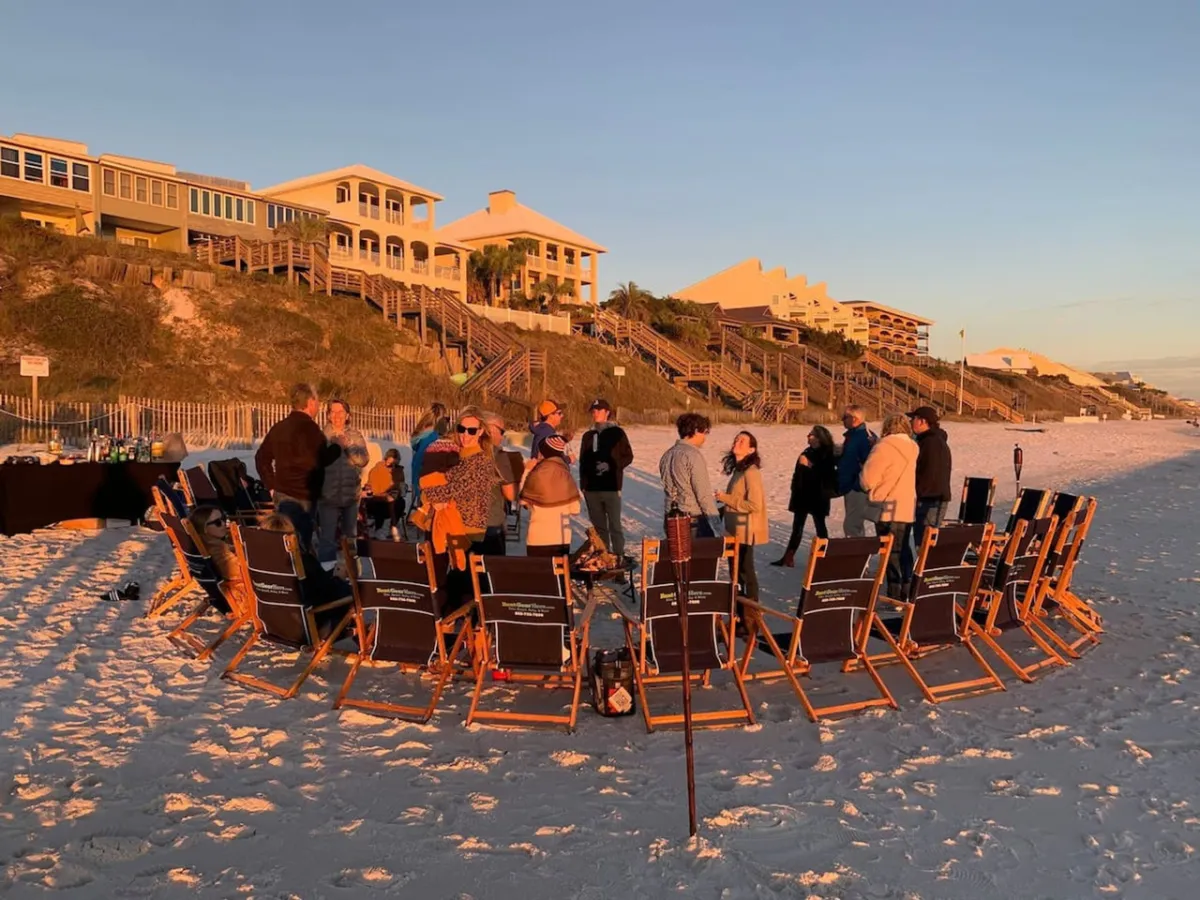 A semi-circle of beach chairs arranged on soft sand, with a small group of people socializing against a backdrop of elevated beach homes. The scene is lively and inviting, with the group enjoying a relaxed gathering, likely during sunset.
