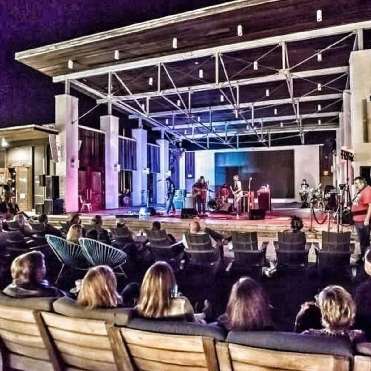 An outdoor concert stage with musicians performing under ambient lighting, surrounded by an audience seated in comfortable chairs. Modern architectural elements and a lively crowd evoke the festive atmosphere of community events.