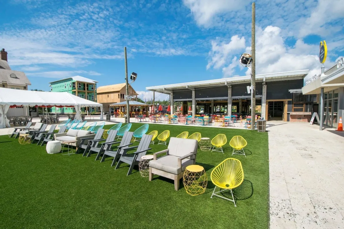 A cheerful exterior scene featuring a row of colorful outdoor chairs, an open pavilion with tables and umbrellas, and a wide green lawn area, suggesting space for community events, outdoor dining, and casual gatherings.