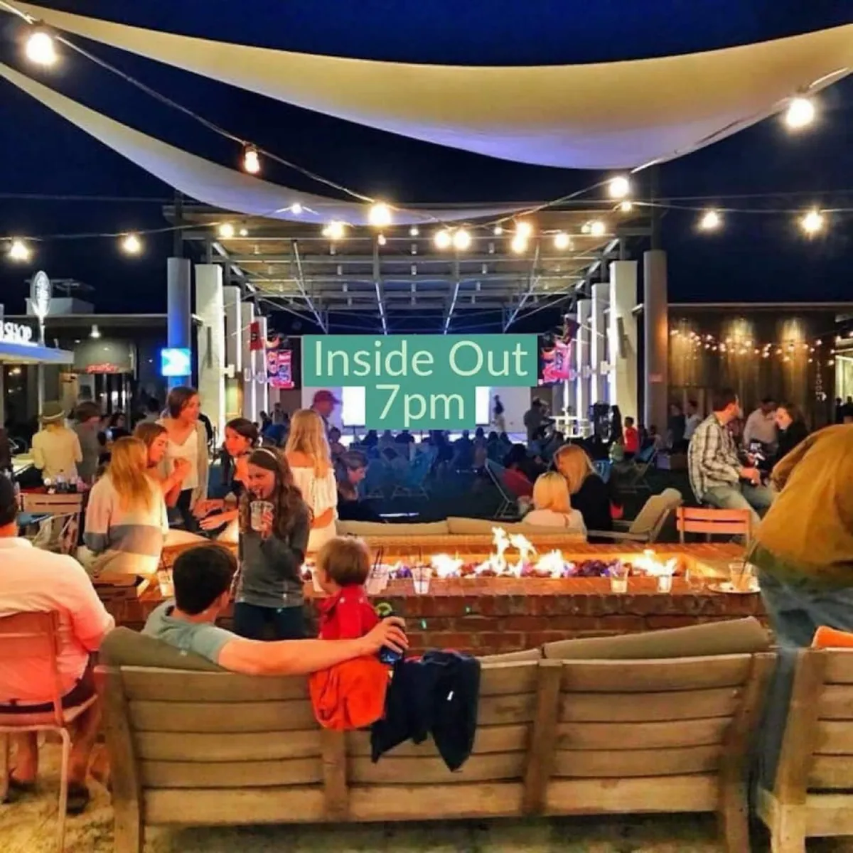 A lively outdoor social scene featuring a central fire pit surrounded by people, string lights overhead, and ample seating. The setting is festive, inviting, and encourages mingling and enjoying community events under the stars.