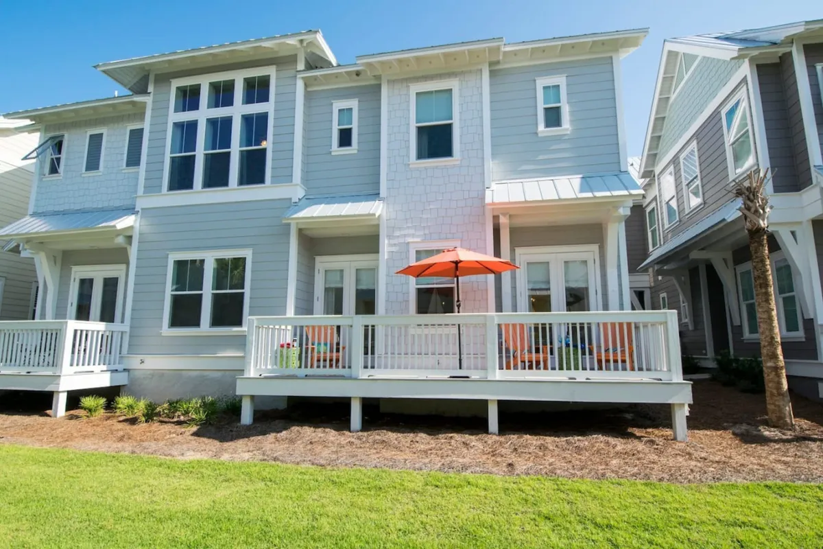 The exterior of a light gray and white townhome with large windows and an elevated deck, accented by an orange patio umbrella. The scene suggests easy access to outdoor amenities and a welcoming, relaxed setting for socializing or family time.