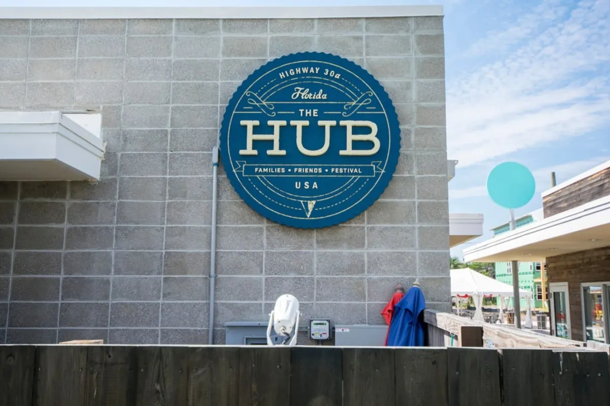 A vibrant scene featuring The HUB sign on a modern building, with outdoor seating areas and people gathering nearby. The setup clearly indicates a lively, social atmosphere, perfect for outdoor dining, events, and community gatherings. The blue circular sign and open layout hint at multiple entertainment and dining options.