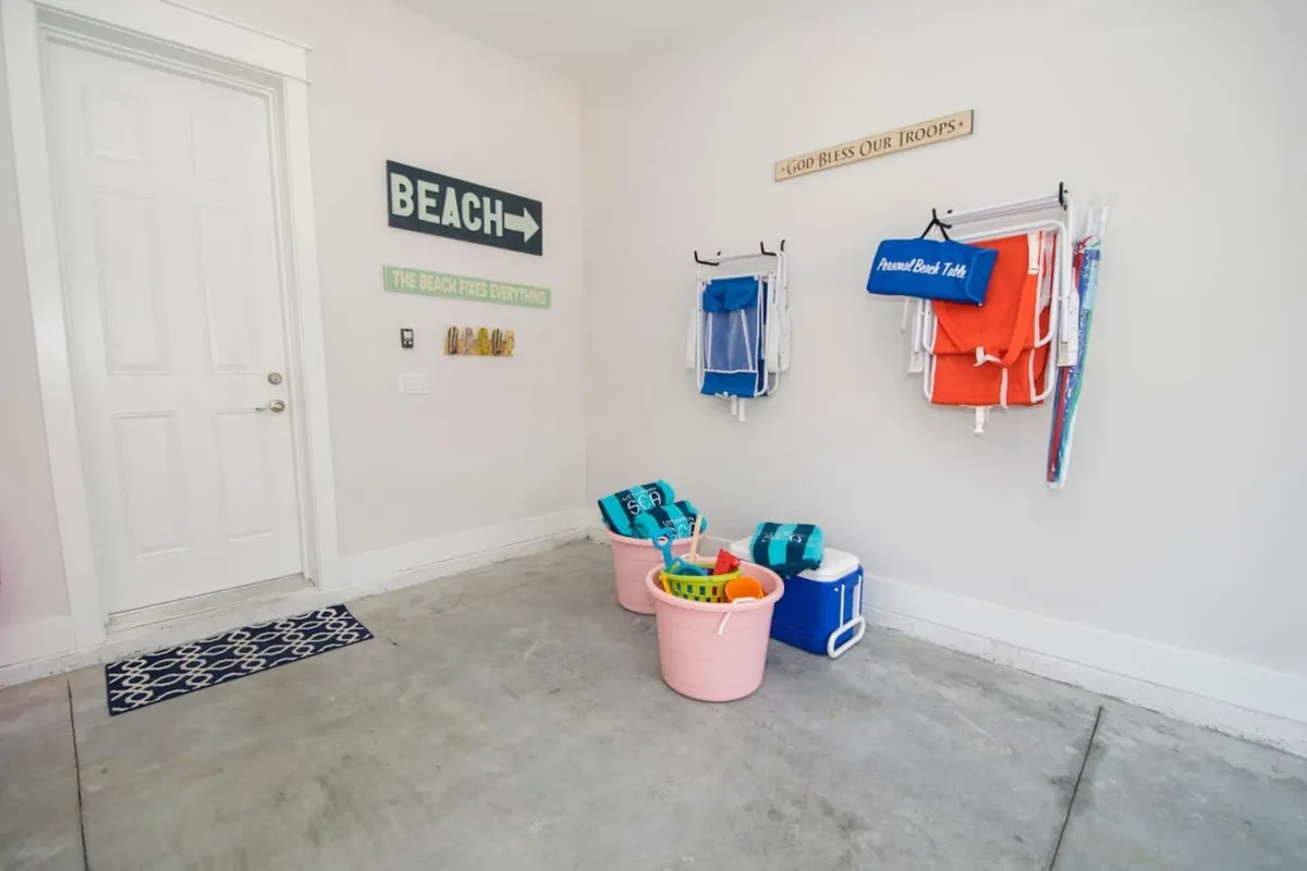 The image features folded beach chairs hung on wall hooks, buckets filled with beach toys and towels, and a cooler, all set against a wall adorned with beach signage. The layout is tidy and inviting, emphasizing ease of access and organization.
