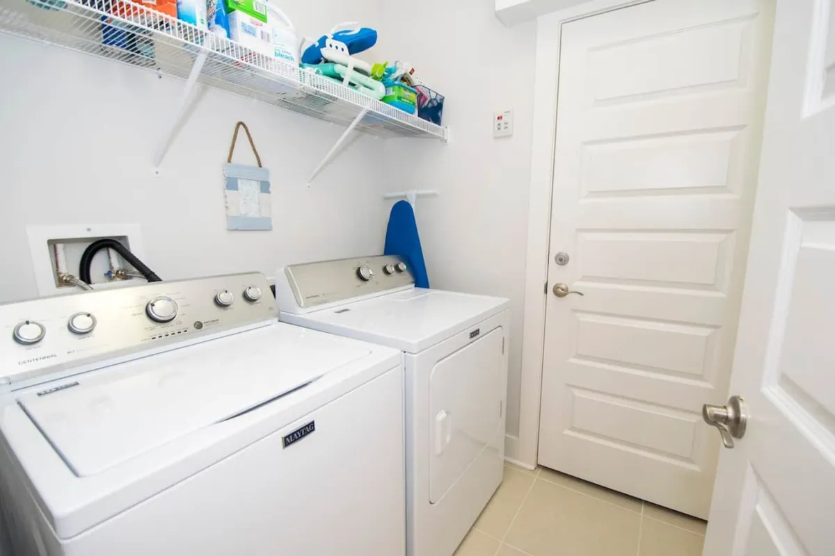 A practical laundry area with a top loading washing machine, front loading dryer, ironing board, and a shelf stocked with laundry supplies. The space is organized and ready for vacationers to keep clothes and towels fresh.