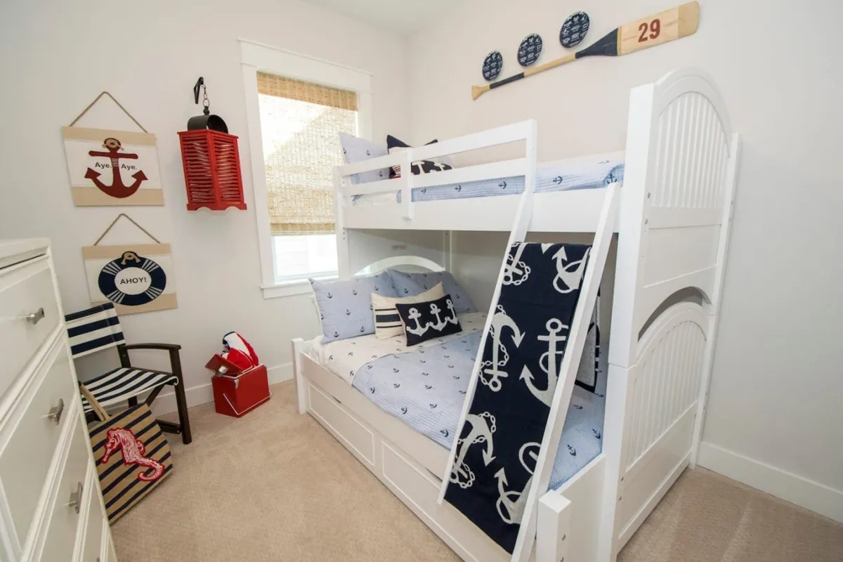 A playful bunk room featuring a white bunk bed with nautical bedding and anchor-themed blanket, red lantern wall decor, nautical wall art, a striped chair, and a toy storage basket. The space is bright, cheerful, and clearly designed with children in mind.