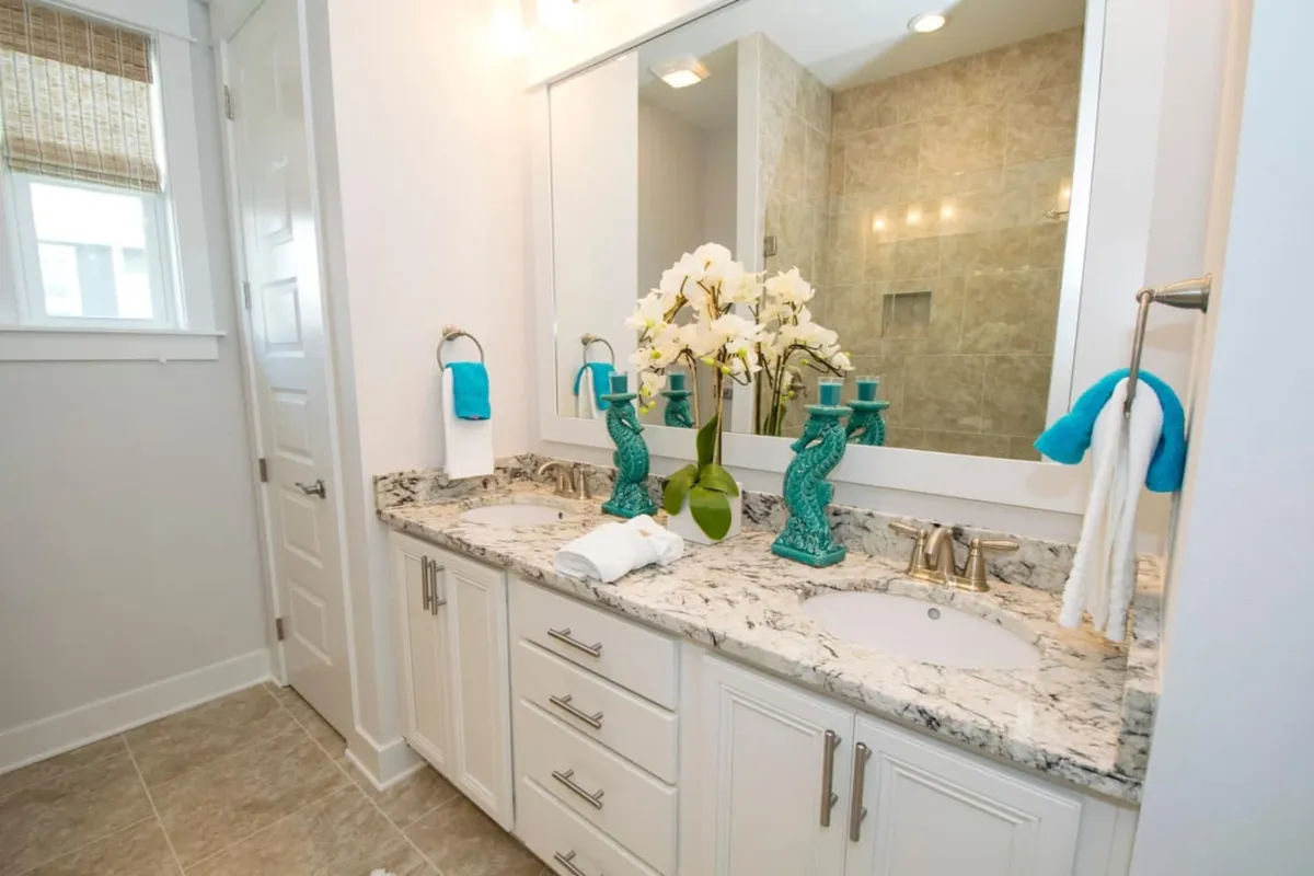 A bright and elegant bathroom with a double sink granite vanity, large mirror, turquoise seahorse candle holders, and a white orchid centerpiece. The scene is airy and spa-like, perfect for morning and evening routines.