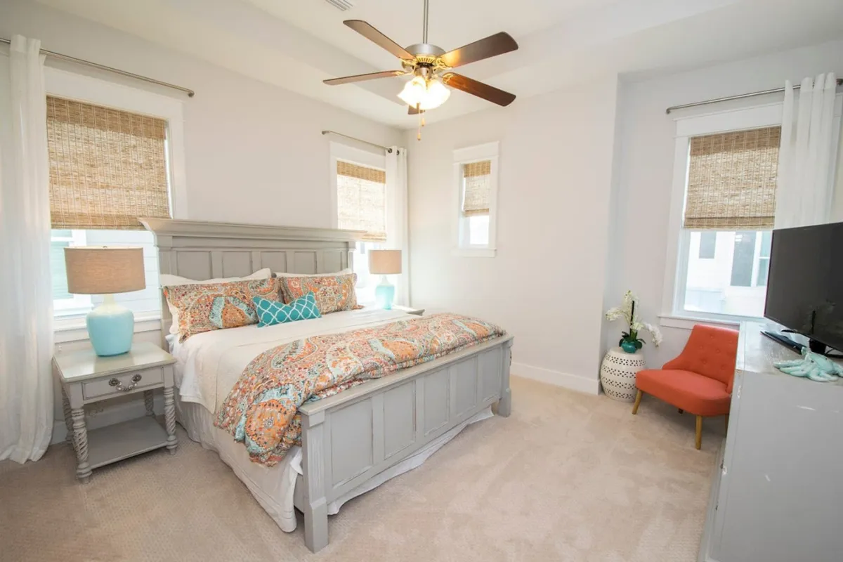 A spacious and serene bedroom with a large grey wooden king bed adorned with patterned bedding, two matching blue bedside lamps, an orange accent chair near a bright window, and a flat-screen TV on a dresser. Natural light fills the room, creating a calm and inviting atmosphere.