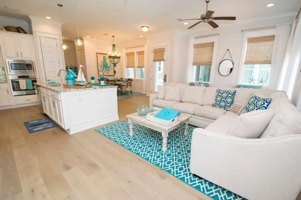 A bright, open-concept living, dining, and kitchen space featuring a large white sectional sofa with patterned blue and green pillows, a central coffee table with decorative items, a white kitchen island with marble countertop and sink, and a dining area highlighted by octopus wall art and a chandelier. Coastal décor and natural light create an inviting, communal atmosphere.