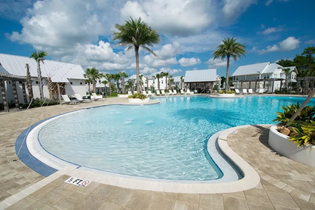A vibrant exterior view of the large zero-entry resort pool surrounded by tall palm trees, with plenty of poolside lounge chairs and shaded cabanas. In the background, a covered pavilion features a grilling area perfect for outdoor dining and gatherings.